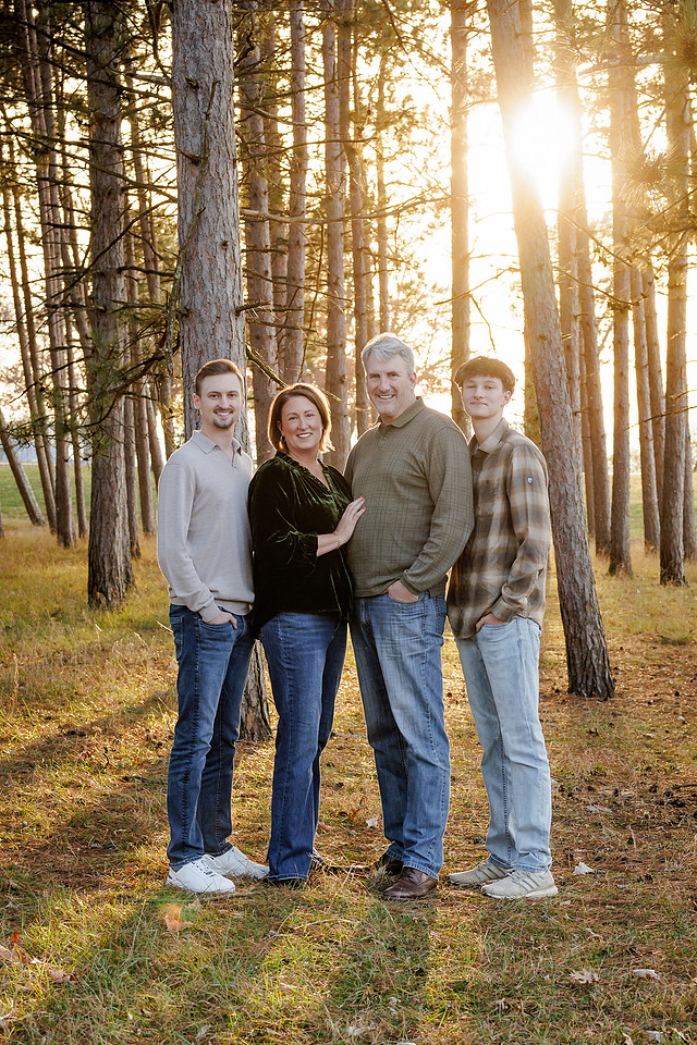 a portrait of a family standing in the woods with the sun filtering through the trees above them